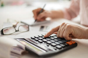 Une main utilise une calculatrice sur des documents, avec des lunettes posées à côté.